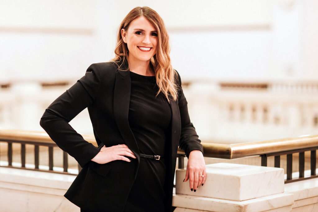 Woman in a black blazer stands with one hand on her hip in a bright interior.