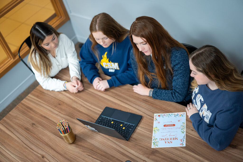 Four students gather around a table looking at a laptop and brochure.