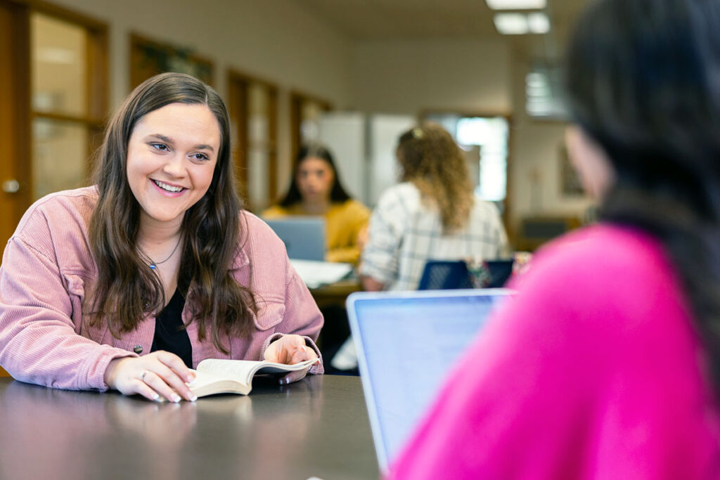Student reads a book while talking with a classmate in a study space.