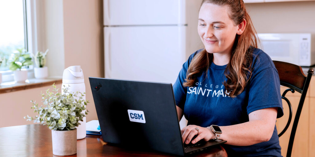 A woman wearing a College of Saint Mary T-shirt works on a laptop at a table.