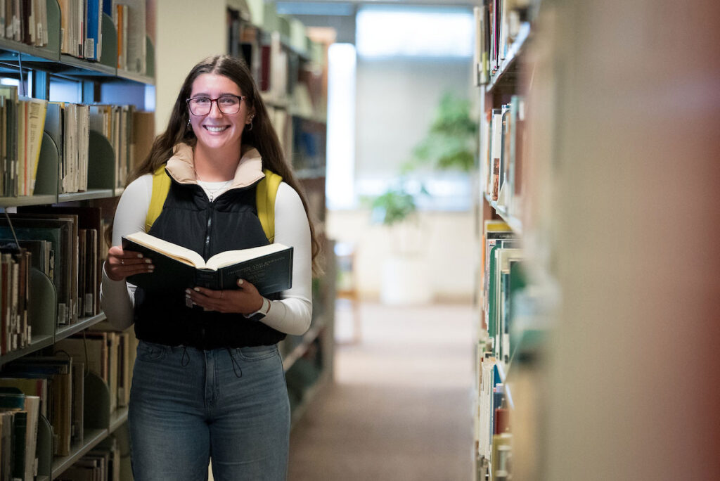 Student smiling with book between library stacks.