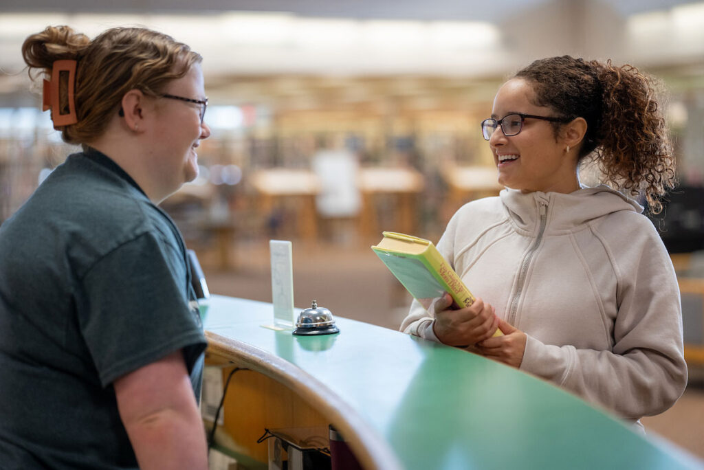 Two students talk at information desk in Library at College of Saint Mary, one holding books.