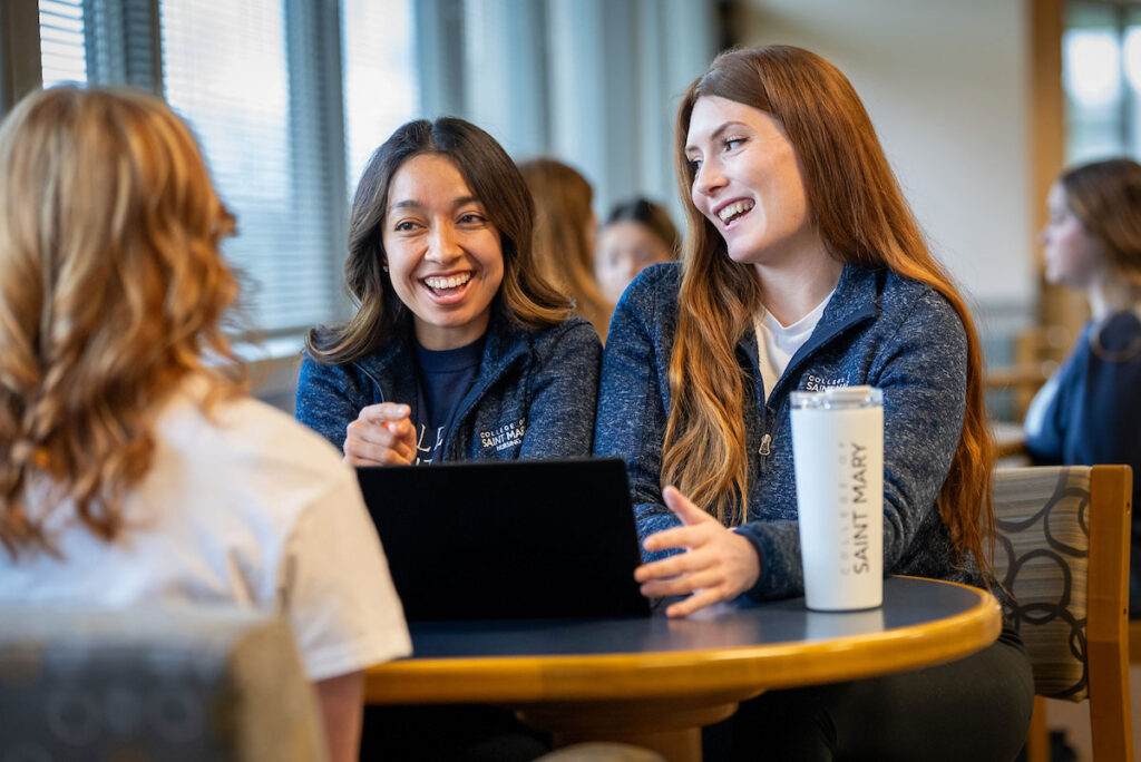 Students talk around a laptop at a table in library with College of Saint Mary water bottle.