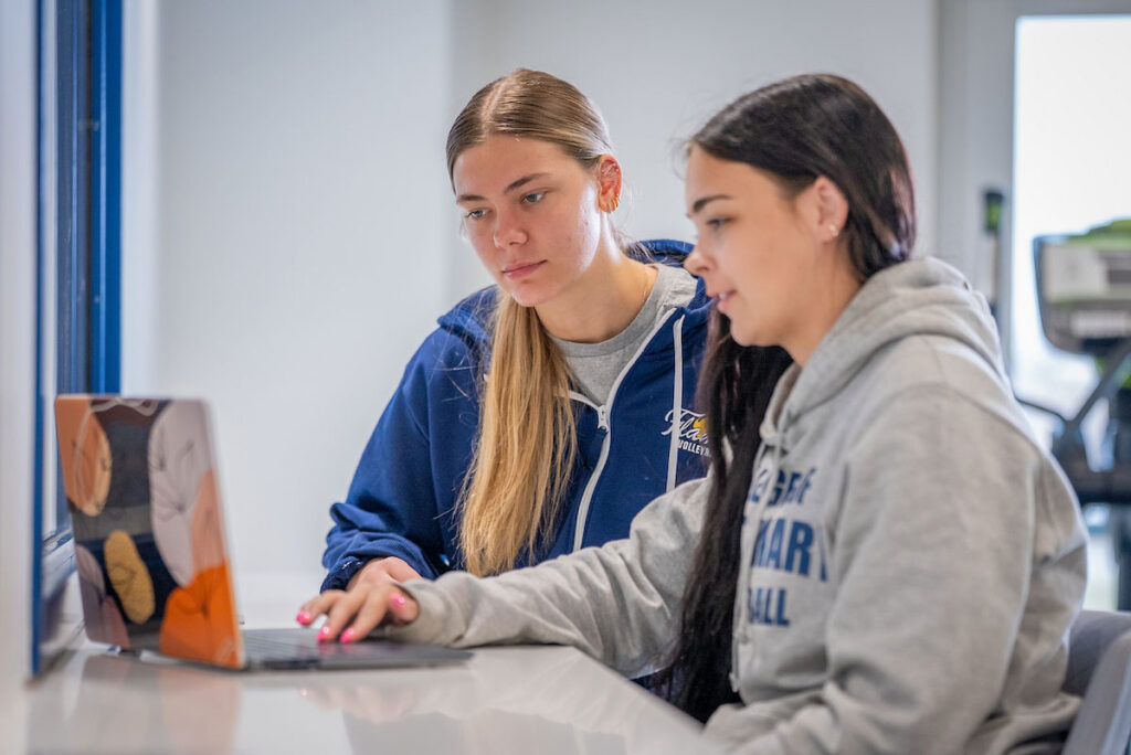 Two students in College of Saint Mary Volleyball sweatshirts looking at laptop together studying.
